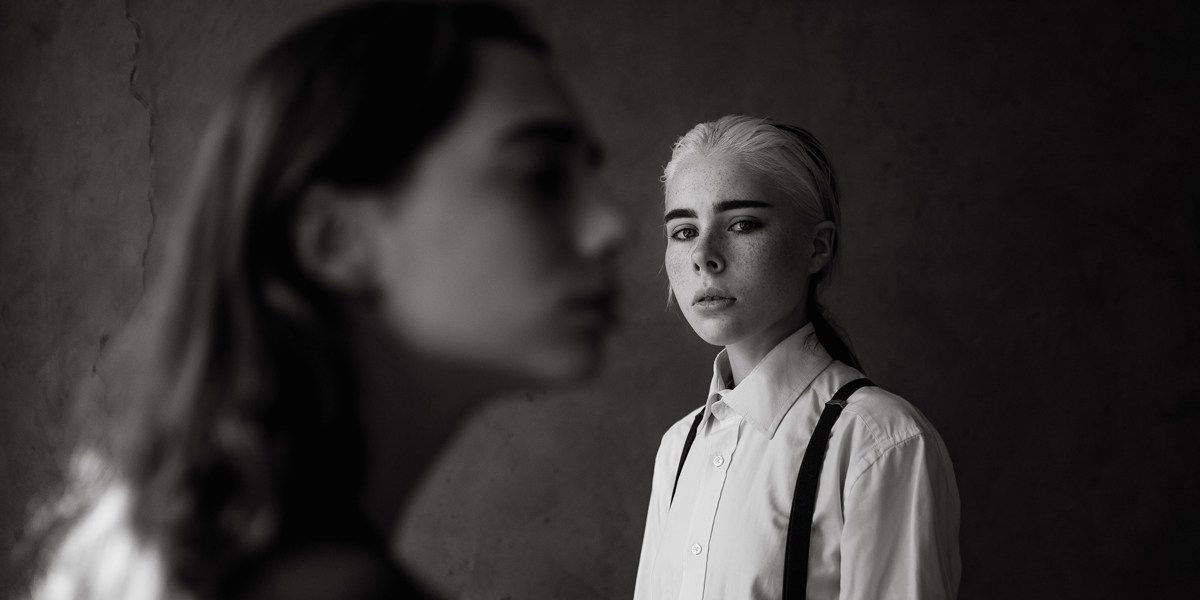 A lonely queer person in a button down and black suspenders, with slicked back blonde hair, looks on longingly in a black and white photo.