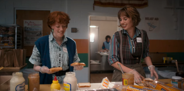 In Stranger Things, Robin and Vickie — two queer white teens in the 1980s, with short hair, Vickie is a redhead and Robin is a brunette — stand next to each other while making Peanut Butter and Jelly sandwiches in a shelter. They are smiling and laughing, and the sunlight shines between them.