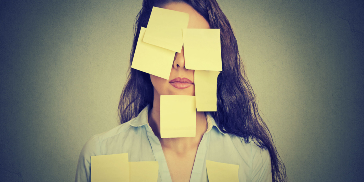 A woman with long brown hair wears light pink lipstick and a light blue button up shirt that's unbuttoned at the top stands against a grey background. Her face and chest are covered in blank yellow Post-it notes.