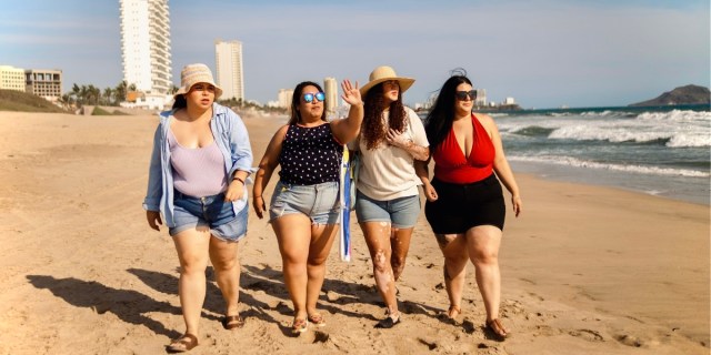A group of four women and people walking together on the beach, left to right: A plus sized person in a purple tank, denim overskirt, white hat, and short shorts; aplus size woman in a black tank top and short shorts and blue sunglasses, a person in white tee, a bucket hat, and shorts; and a plus sized woman in a low cut red one piece bathing suit and black shorts.