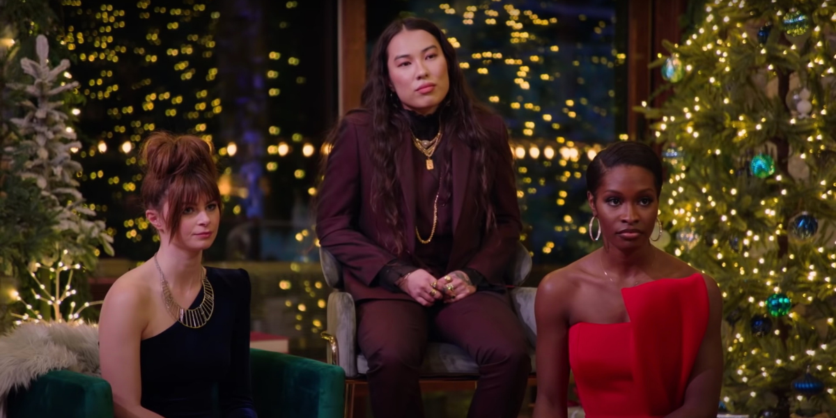 Rachel, Hina, and Krissi sit in the 12 Dates of Christmas cabin surrounded by Christmas light-covered trees during the gift ceremony. Rachel is wearing a black dress. Krissi is wearing a red dress. Hina is sitting on a chair that's elevated and they're wearing a dark purple suit.