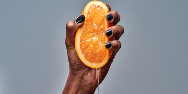Against a grey background, a Black woman's hand squeezes half of an orange. The juice drips down her wrist. She is wearing black nail polish.