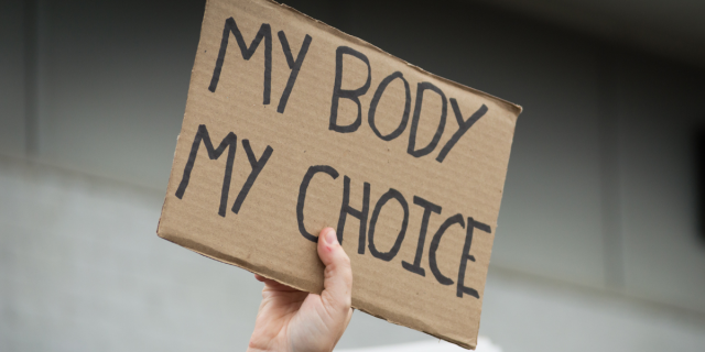 Against a white tiled background, a white person's hand holds up a cardboard sign that reads, "MY BODY MY CHOICE."
