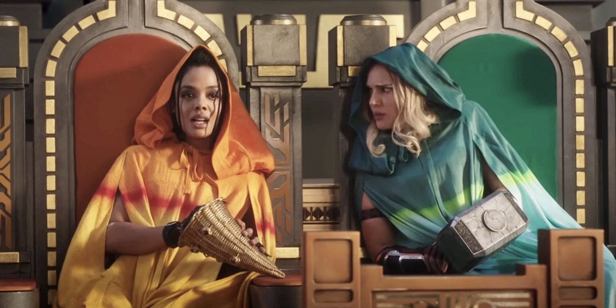 In Thor: Love and Thunder, Valkyrie, who's now gay on screen, sits next to Jane Foster. Valkyrie is played by Tessa Thompson, Jane is played by Natalie Portman. The two actresses are in matching gold thrones, one with red velvet seats and one with green. Valkyrie is in a gold robe with a hood, and Jane is in a green robe with a hood. Jane is leaning in to talk with Valkyrie.
