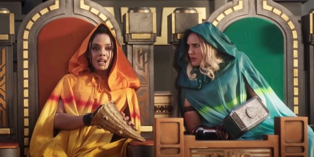 In Thor: Love and Thunder, Valkyrie, who's now gay on screen, sits next to Jane Foster. Valkyrie is played by Tessa Thompson, Jane is played by Natalie Portman. The two actresses are in matching gold thrones, one with red velvet seats and one with green. Valkyrie is in a gold robe with a hood, and Jane is in a green robe with a hood. Jane is leaning in to talk with Valkyrie.