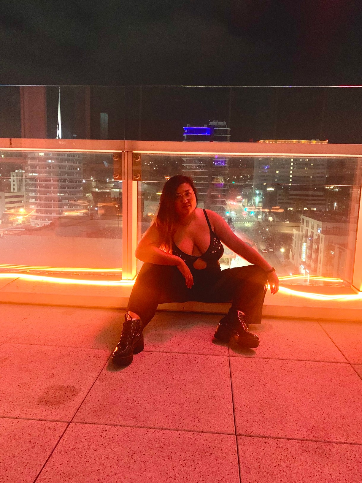 Em is a mixed-race Asian person who is sitting on what looks like a bridge overlooking a city. They are wearing a cool, all black outfit and leaning on one knee.