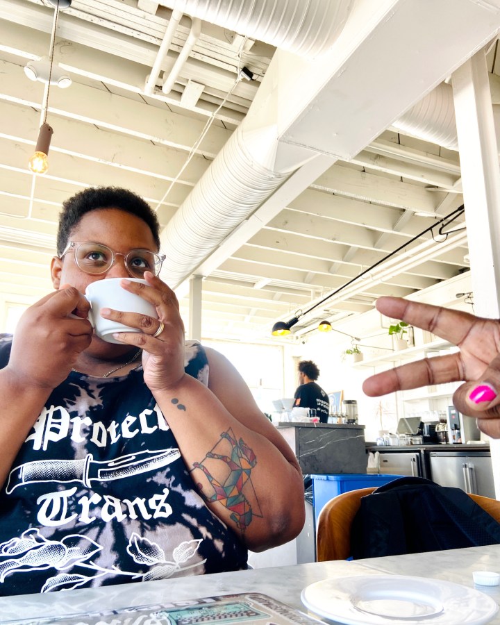 shea, a Black queer person wearing a PROTECT TRANS KIDS tshirt sips a cup of coffee with a friend