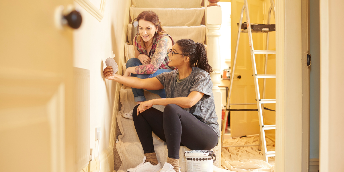 two queer women sit on the stairs comparing paint swatches. a ladder and drop cloth are visible in the background.