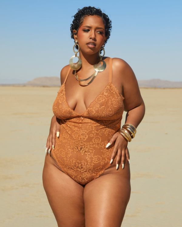 A plus size black woman in a Lingere teddy that matches her warm almond skin tone.  She has short cropped hair and white nails.  She is on the beach.
