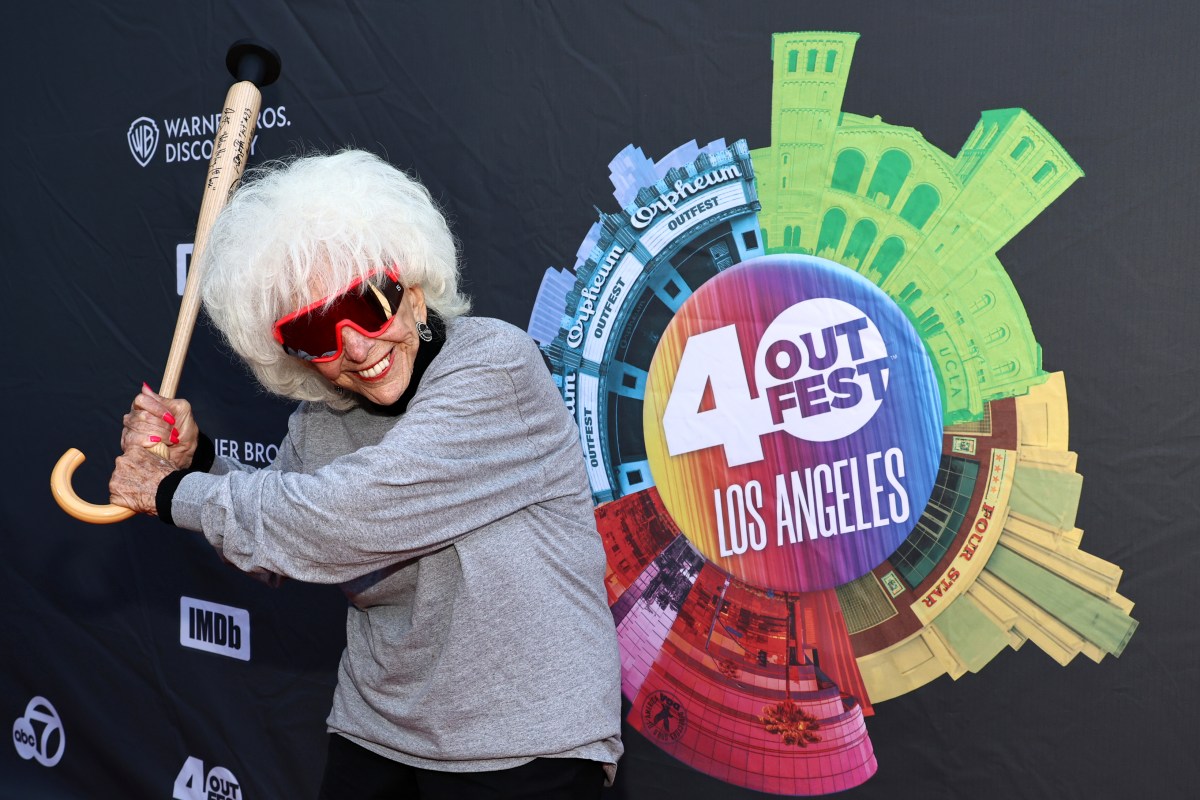 an elderly white woman in sunglasses holding up a cane in the shape of a baseball bat. Outfest 40th anniversay signage behind her