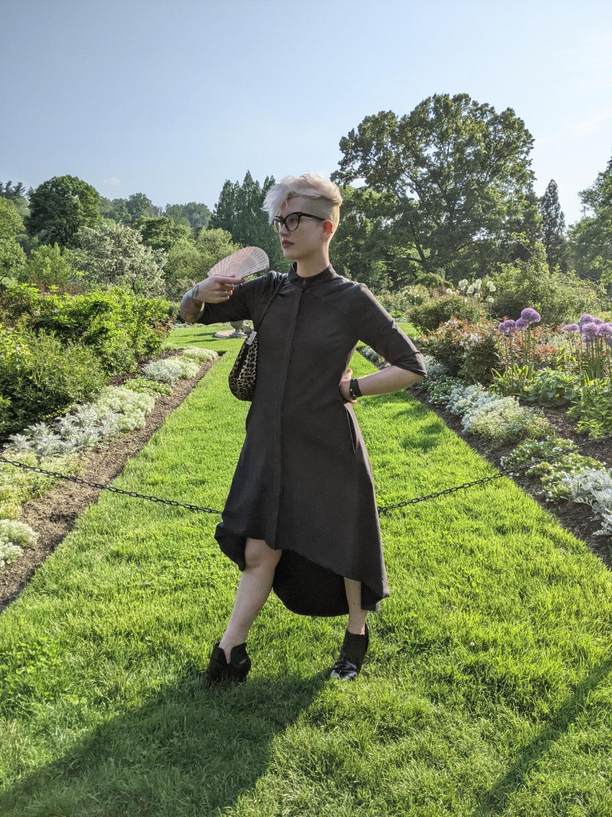 Nicole, a white genderqueer human, stands in a well manicured botanic garden in a dress, in a hot mean pose. It is also literally 99 degrees where they are. they have shaved sides and bleached hair.