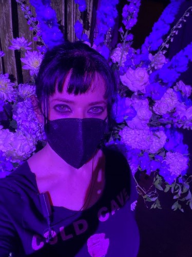 Stef, a white woman, poses with a mask on in a room with purple ighting in front of a bouquet of flowers. Stef has black hair pulled back.