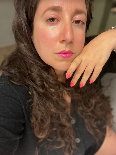 Vanessa, a white woman, poses with her hand to the side of her face. She is gazing hotly at the camera. Her nails are painted and she has pink lipstick on. Vanessa has long brown hair.