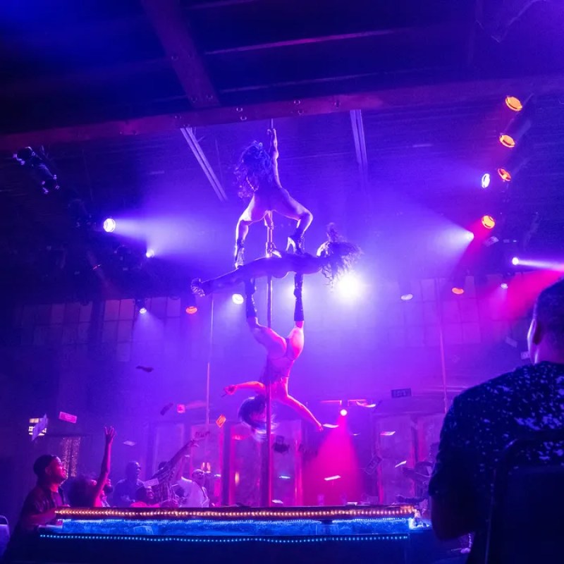 In the show P-Valley, two dancers work together on a pole in front of a large crowd underneath blue and pink lights