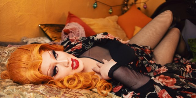 Jinkx Monsoon lies in bed wearing a big ginger wig. She has on a black dress with reddish pink flowers, see through sleeves, and fishnet stockings.