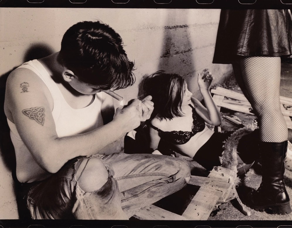 A black and white image shows a person in a white tank top and ripped jeans with short brown hair. They have a Celtic design tattooed on their arm, as well as a smaller tattoo of a cartoon boy. They lean over to light a cigarette. Beside them, a woman with shoulder length brown hair with a blonde highlight lounges in a black lace bra and skirt. The black leather skirt and fishnet-clad legs of a third person are visible to the right. Scraps of wood surround them.
