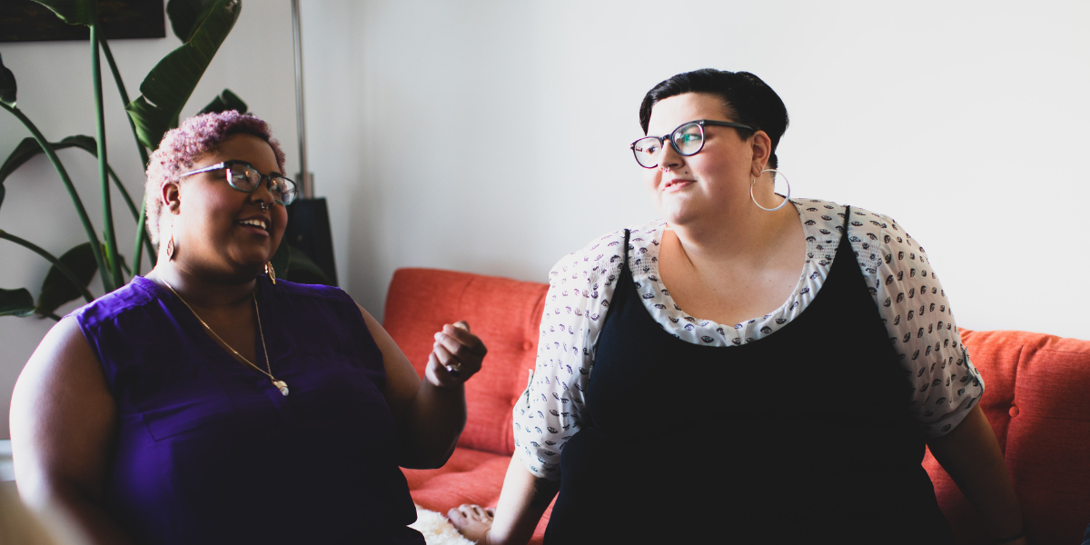 Two people having a conversation in a living room having a conversation. The person on the left is Black with short pink curly hair. The person on the right is white with short black hair. They are smiling at one another.