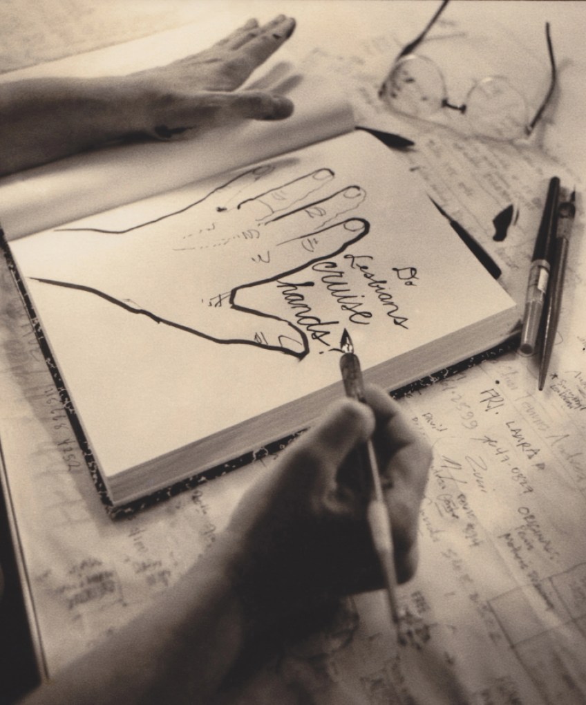 A black and white photos shows a sketchbook featuring a line drawing of a hand. Next to the hand are the words, "Do lesbians cruise hands?" in cursive. The hands of the artist are visible on either side of the sketchbook. Their right hand holds a pen, and the first finger on their left hand is dotted with ink. Paper with scribbled names and numbers is underneath the sketchbook. The artist's glasses sit above the sketchbook.