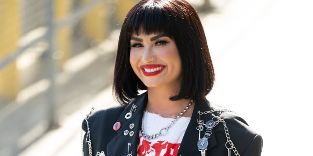 Demi Lovato has on bright red lipstick and a bob haircut with bangs, they are wearing a black leather motorcycle jacket and standing outside on a bright, sunny day.