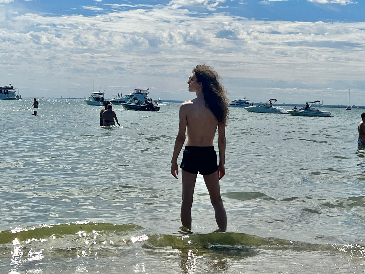 Drew stands in a body of water as seen from behind. She is topless and wearing a pair of black shorts and sunglasses. She is seen from behind with her long curly brown hair blowing in the wind. There are multiple boats and people swimming in the background. Drew is a white woman.