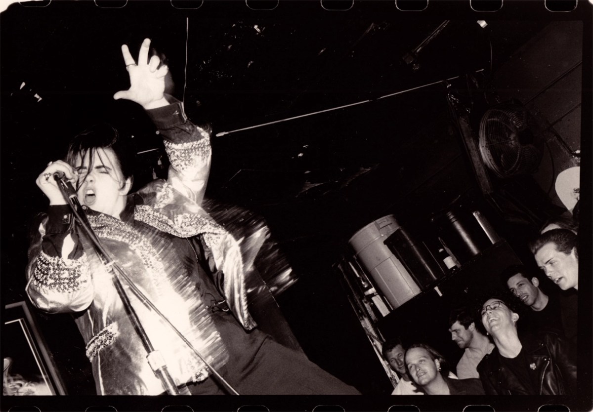 A black and white image shows a drag king dressed like Elvis. The king wears a gold, bedazzled jacket, a black shirt and black pants. He holds a microphone on a stand and throws one of his arms into the air while singing. His black hair falls into his face. Audience members smile in the bottom right corner of the image below the stage.