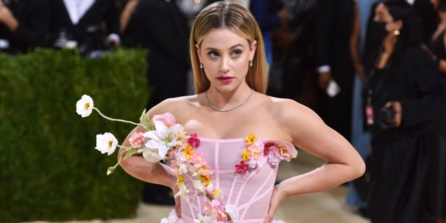 Actor Lili Reinhart, who will be starring in a queer period drama soon, is walking the MET red carpet in a pink dress with a corset and flowers. She's posed with her hands behind her back.