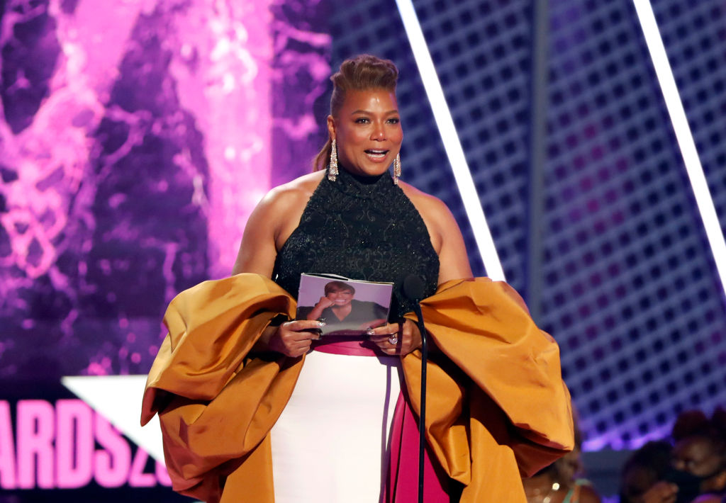 Queen Latifah accepts the Lifetime Achievement BET Award onstage at the BET Awards 2021 at Microsoft Theater on June 27, 2021 in Los Angeles, California. She is wearing a big yellow billowing shawl type accent piece over a glittery black top and smiling brightly. Queen Latifah is a Black woman with brown hair back in an updo style and long earrings.