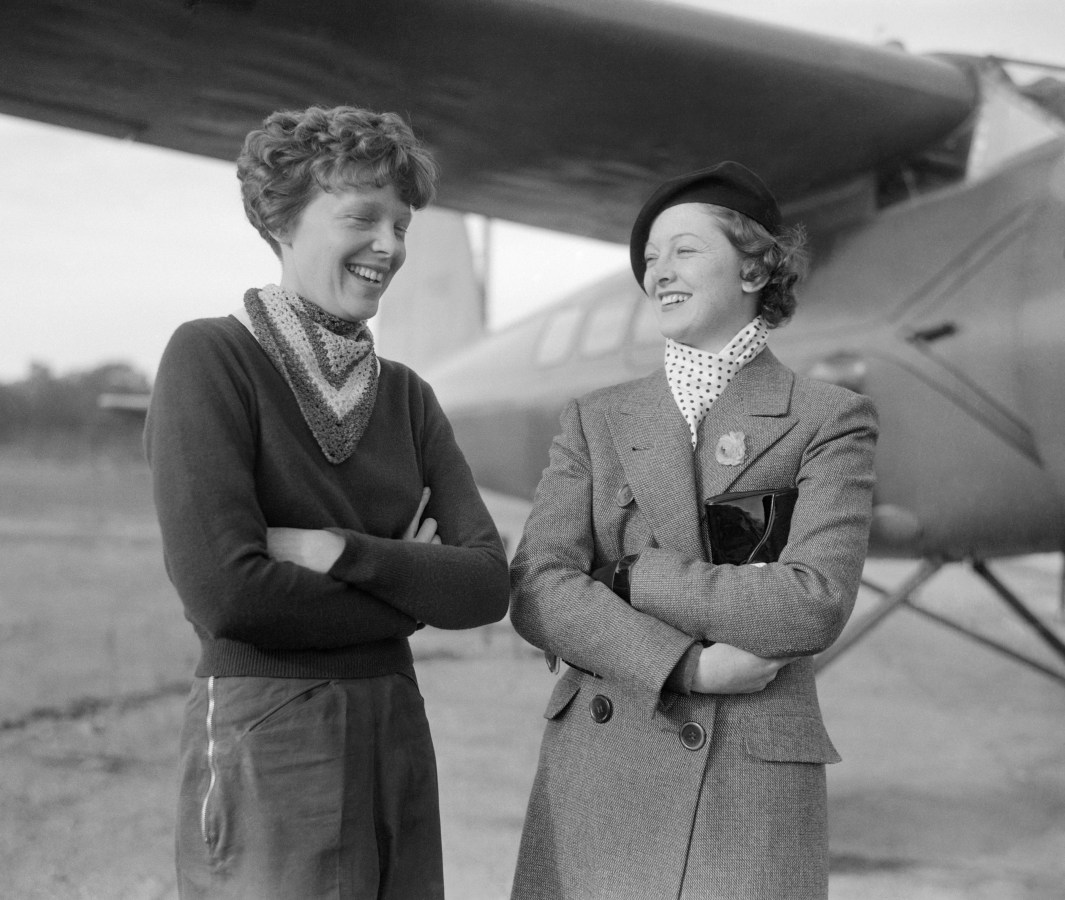 Amelia Earhart and Myrna Loy stand next to each other in front of a plane. They both have their arms crossed and are both chuckling.