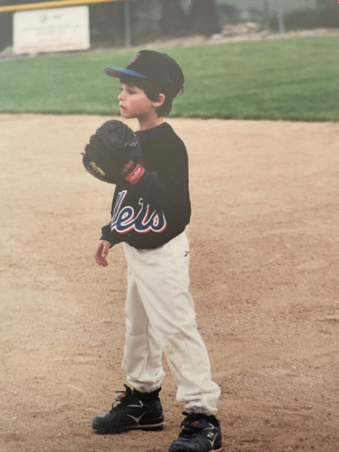 The writer, Drew, as a young child wears a Mets jersey, baseball hat, and baseball glove while standing in a baseball diamond.