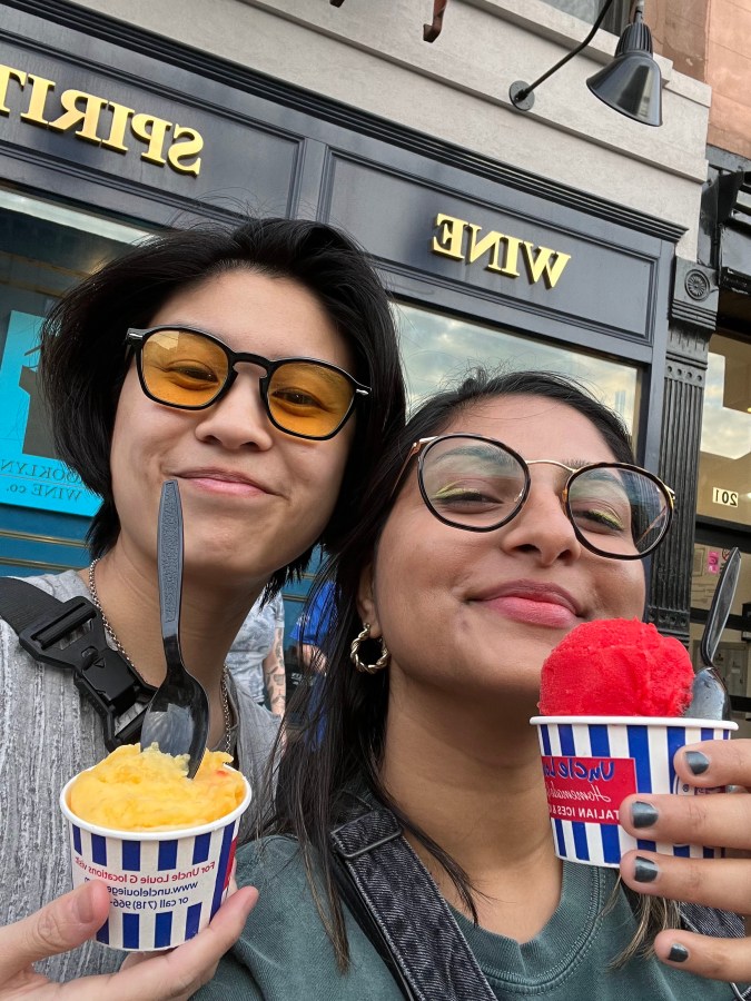 Gerrie, a Chinese-Filipino nonbinary person has a short haircut and black hair and is wearing yellow tinted sunglasses and smiling. Next to them, Ashni, a gender averse South Asian person wearing prescription glasses is also smiling. They are both holding up italian ices, one yellow, one red.