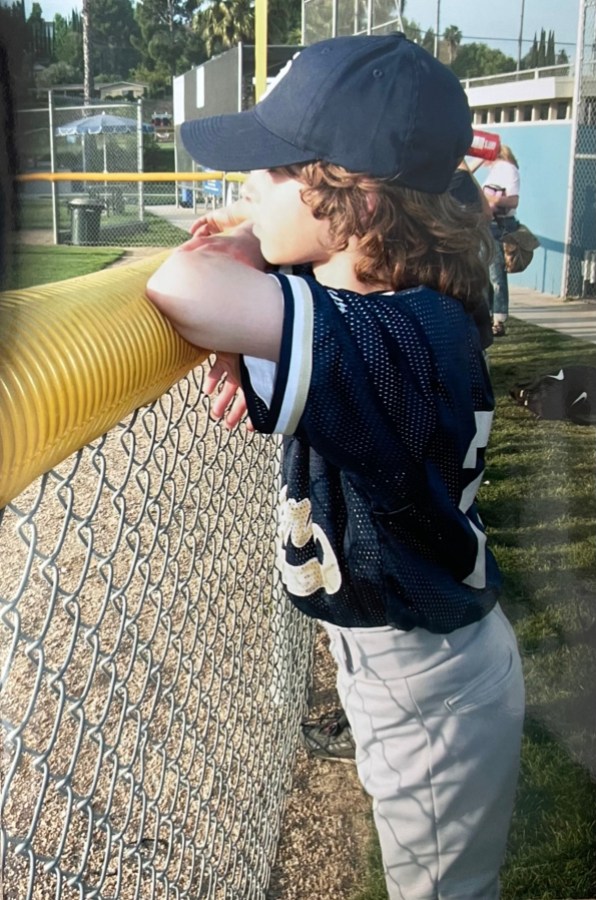 The writer, Drew, as a young child wears a baseball hat and baseball uniform while propping her arms up on the side of a fence next to a baseball diamond.