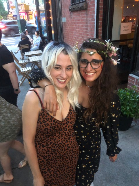 A woman dressed in costume as a young Nat from Yellowjackets and Kayla Kumari Upadhyaya wearing an antler crown and floral dress with fake blood on her mouth, dressed as Lottie.