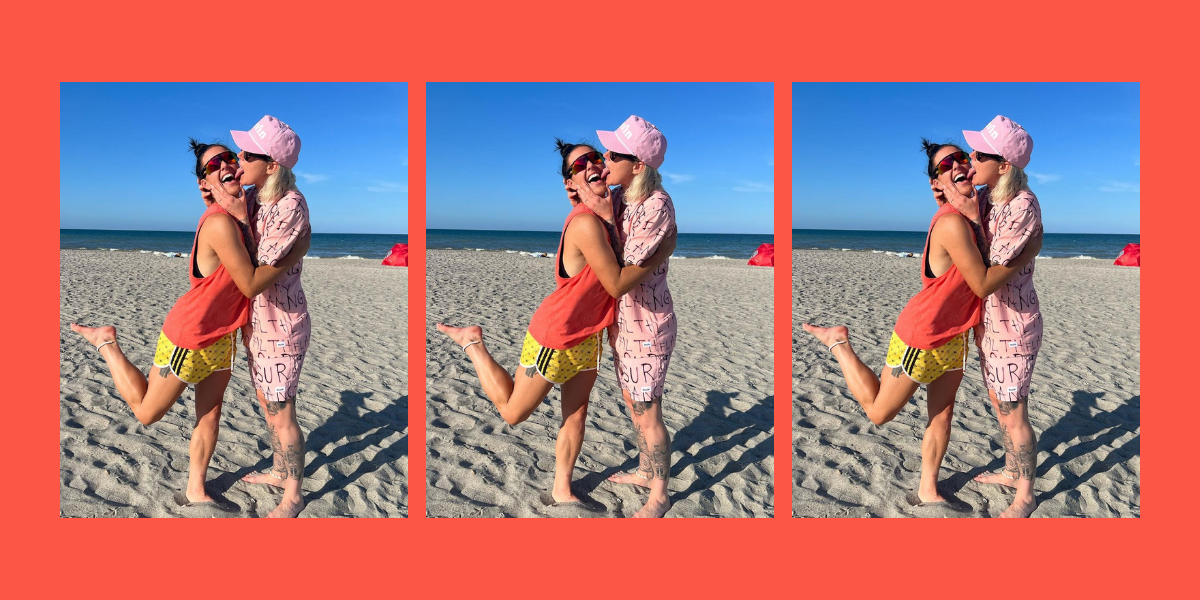 Ashlyn Harris wears a pink shirt and pink shorts and pink hat on the beach while holding and licking the face of Ali Krieger who is wearing a red rank and yellow shorts.