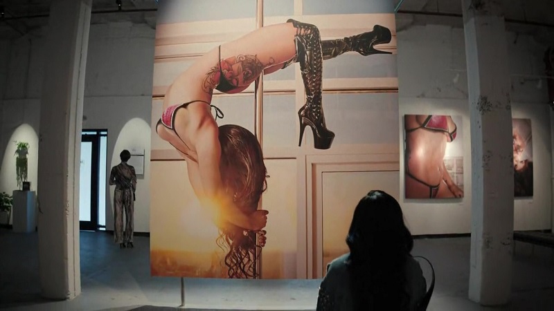 Mercedes sits, wearing a denim jacket, staring at a large photo of her. In the photo, she's wearing her floss and knee high stileto boots, hanging upside down on the pole.