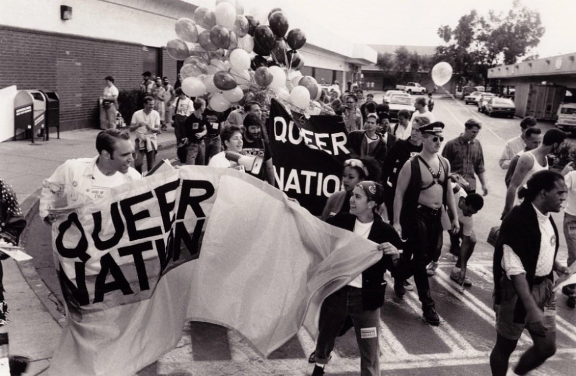 A black and white image shows a crowd of queer people in the street at a protest. Two people on the left hold a fabric sign that reads, "QUEER NATION" in black letters. Behind them, two more people hold a black fabric sign that reads, "QUEER NATION" in white letters. A large bundle of balloons is in the background. All of the participants are wearing typical street clothes, but one man is wearing a leather vest, a leather harness, a leather cap, leather pants and sunglasses. 
