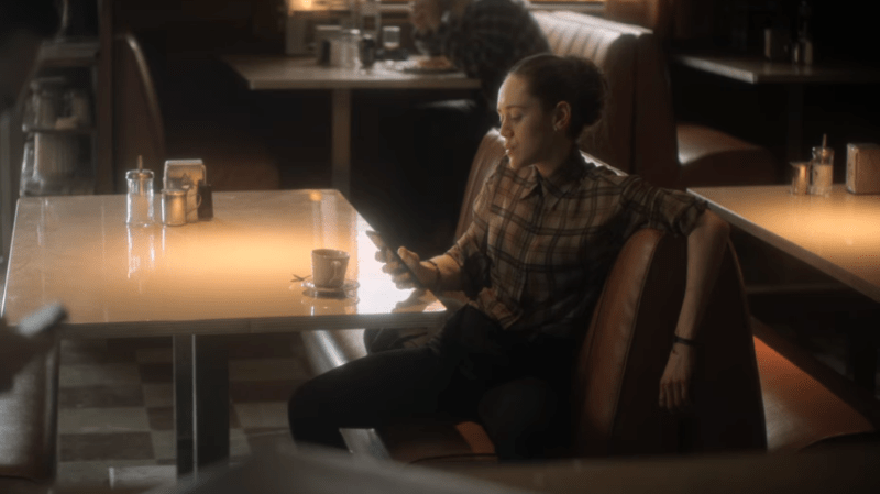 The Sandman: Judy is sitting gayly in a diner booth while scrolling on her phone, wearing a yellow and black plaid shirt buttoned all the way up to the neck