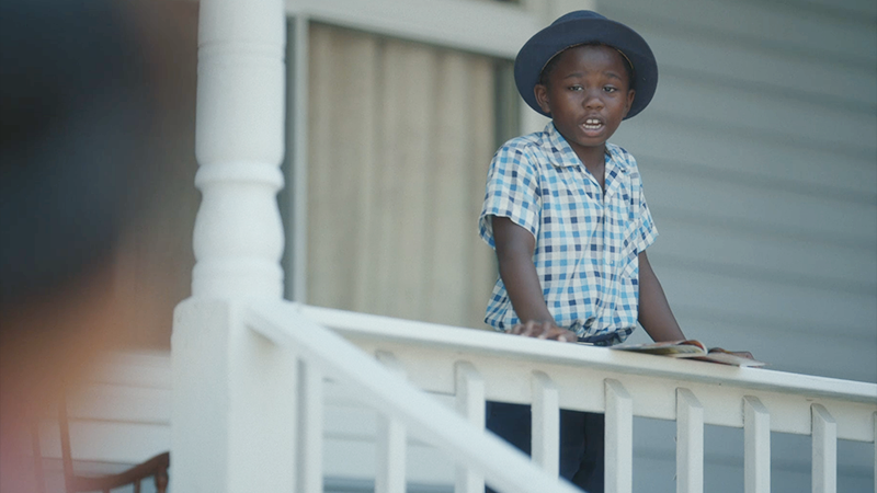 A young Black boy in a hat on a porch. 
