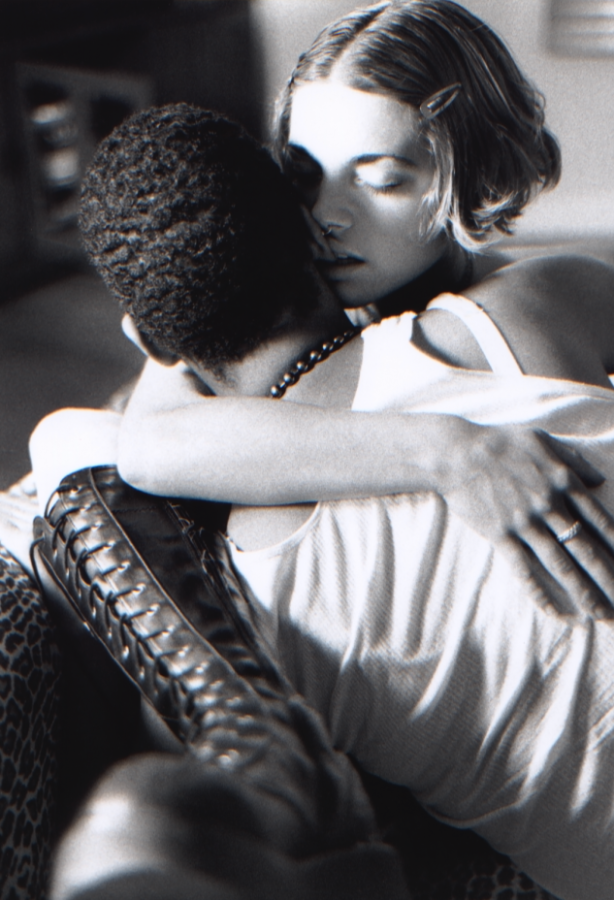 A black and white image shows a white woman with short bob haircut wearing black combat boots. She is embracing a Black woman, who has short hair and wears a white tank top and a black beaded necklace. The two are seated on what looks like a leopard print couch or chair. The Black woman faces away from the camera. 