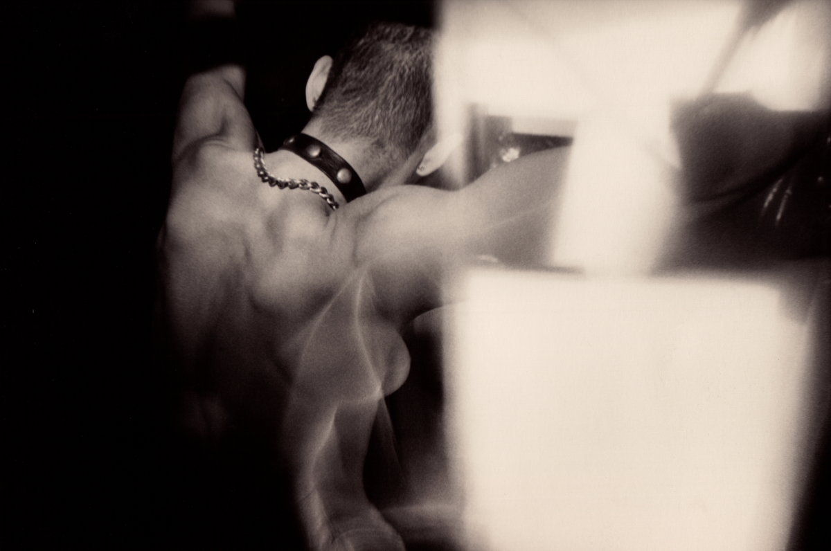 A black and white image shows a shirtless person with a buzz cut wearing a black studded collar and a chain around their neck. Their arms are in the air and hey are facing away from the camera. Their muscular back and the side of their right breast are visible. Flashes of light are on the right.