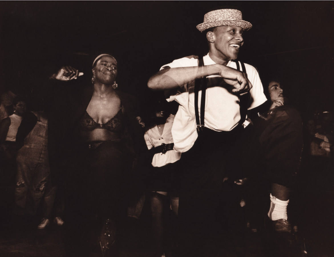 A black and white image shows a Black woman wearing a headband, an open black blazer with a black lace bra underneath and black pants dancing. Beside her is a Black masculine person, who is wearing a brimmed hat, a white T-shirt, suspenders, black pants, white socks and dress shoes. They are also dancing and smiling. More dancers are visible in the background.