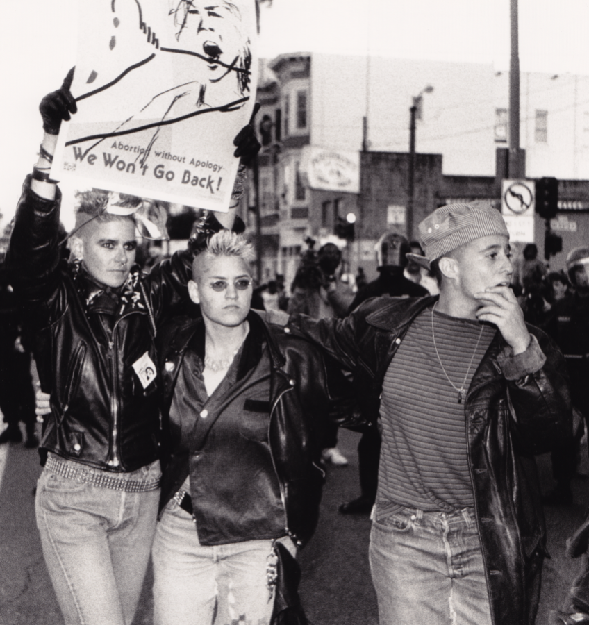 A black and white image shows three women standing in the street at a protest. On the left, a woman with short, spikey blonde hair that's shaved on the sides wears black eyeliner, a black leather jacket, jeans, a studded belt, and black leather gloves. She holds a sign with an image of screaming woman and coat hanger that reads, "Abortion without apology. We Won't Go Back!" In the center, there is a woman with short, spikey blonde hair that's shaved on the sides. She wears a black button up shirt, a black leather jacket, jeans and sunglasses. On the right, there is a woman wearing a striped backwards hat, a striped shirt, a black leather trench coat, jeans and a necklace. She looks off to the right and touches her hand to her chin while wrapping her arm around the middle woman's shoulders. Behind them, more protestors and cops in riot gear are visible.