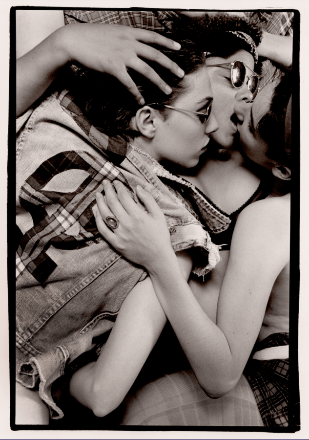 A black and white image shows three women lying on top of each other in a pile with their arms wrapped around each other. Two of the women — one with a bandanna around her forehead and sunglasses and one wearing a headband and no shirt — are kissing. The third woman, who wears sunglasses and a denim vest with a plaid peace sign/Venus symbol on the back, leans in towards the mouths of the kissing women.