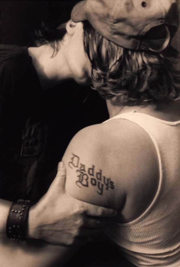 A black and white image shows the back of a person with chin-length, wavy brown hair wearing a baseball cap and a white tank top. They have the words, "Daddy's Boy" tattooed on their left arm. Another person whose face isn't visible wears a black shirt and a black leather wristband. They grip the first person's arm and lean in to kiss them.