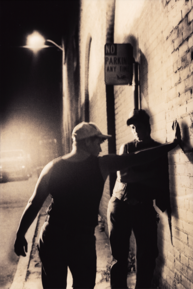  A black and white image shows the silhouette of a masculine person wearing a baseball cap and jacket leaning against a brick wall in front of a "No Parking Any Time" sign. Another masculine person, whose back is to the camera, stretches out their arm to press their right hand against the brick wall. They wear a baseball cap and a black tank top. A street lamp is visible in the background.