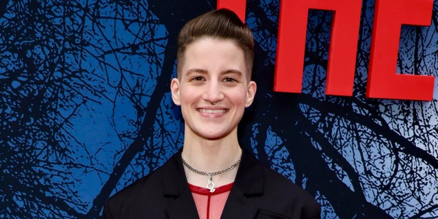 Theo Germaine, a white nonbinary trans person with short brown hair, is smiling on the They/Them red carped (there's a bright red They/Them sign behind them). They have a pink shirt, a metal choker, a black jacket, and a nose ring.