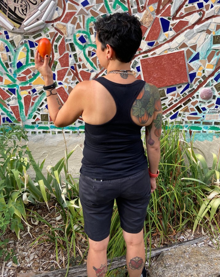 Tracy is seen from behind in the community garden holding up a tomato. Tracy is an Asian woman with a bunch of tattoos and dark hair.
