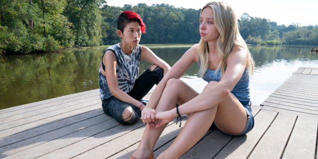 A still from They/Them in which Veronica [Monique Kim], a bisexual Asian woman with dyed red and black hair buzzed short on one side and Kim [Anna Lore] a white lesbian with long blonde hair, sit on a dock on a lake at a gay conversion camp. They have looks of deep concern and worry on their faces.