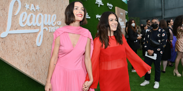 Jodi Balfour wears a pink dress and holds hands with Abbi Jacobson, who she is engaged to and who is wearing a red dress at the League of Their Own premiere red carpet.