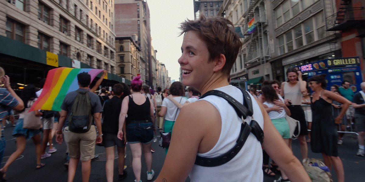 Leo Baker wears a white tank and a leather harness while walking in a Pride parade