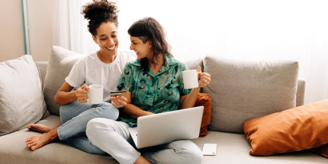 two queer women one who is Black with her hair in a top puff and one who is white with a shaved side and long brown hair laugh together over coffee and computers